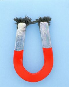 Ein Dauermagnet in Hufeisenform, auf dessen einem Pol ein S, auf dem anderen ein N steht und an denen sich an den Spitzen Eisenspäne gesammelt haben. der untere Teil ist rot.